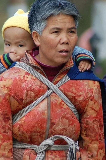 An older woman stands upright and purposeful, her expression focused as she moves through a public space. Across her chest, a simple cloth wrap is crossed in an “X” and tied firmly at her waist, anchoring the child on her back with practiced confiden