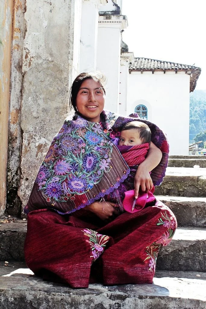 Seated on stone steps beside a stuccoed wall, a young mother gazes warmly at the camera as sunlight catches the vivid embroidery of her garments. Her baby is nestled against her chest in a richly woven wrap of deep pink, tucked into the folds of a tr