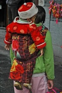 A toddler sleeps peacefully on their mother’s back, nestled into the familiar warmth of a Chinese bei bei — a traditional quilted carrier still widely used across China today. The carrier’s bright floral fabric, complete with bold red roses and styli