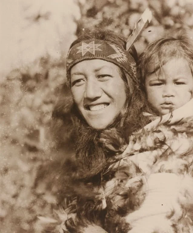 With sunlight soft on their faces, this radiant image captures a Māori mother and child carried close in a feathered korowai. Her smile is wide and unguarded, framed by long flowing hair and a woven headband adorned with a stylised star motif. A feat