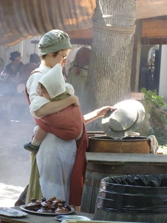 A quiet, tender moment captured amid the busyness of daily tasks. This reenactor — dressed in historical attire evocative of early European settler or colonial domestic life — lifts a cooking pot while her child sleeps soundly on her front. The baby’