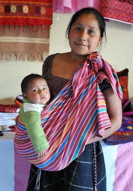A young mother carries her baby in a traditional striped rebozo, tied securely over one shoulder in a sling-style carry. The cloth is vibrant, woven in shades of pink, red, turquoise, and white — echoing the rich textile heritage of southern Mexico,