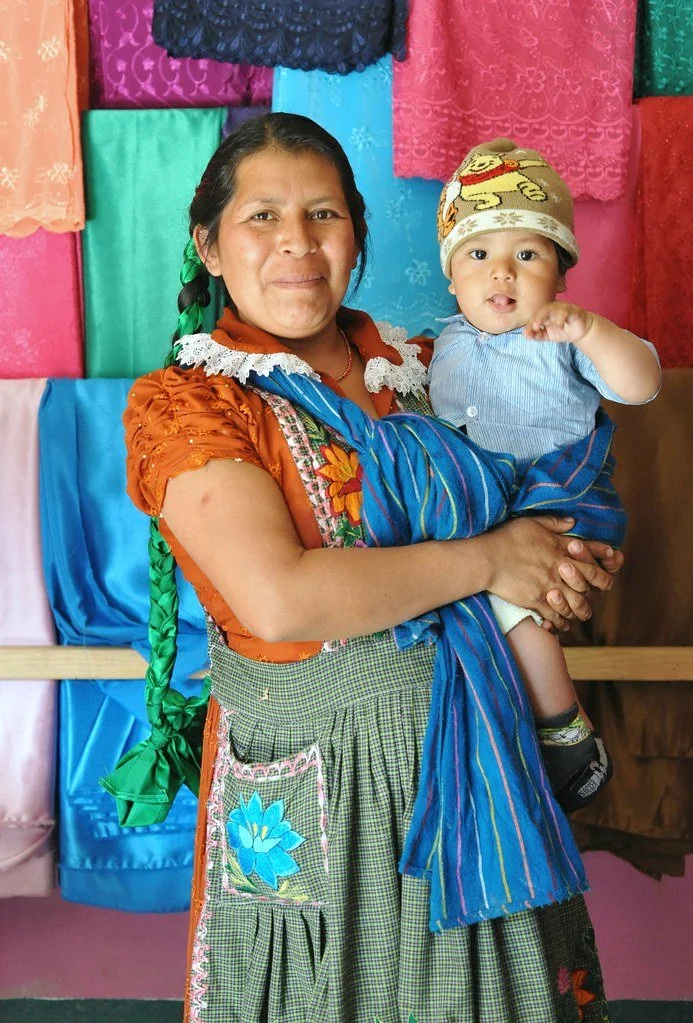 A mother stands proudly in front of a vivid backdrop of folded textiles, her baby cradled comfortably in her arms. She wears a green gingham apron over a traditional embroidered blouse in warm earth tones, her long braid wrapped in bright green ribbo