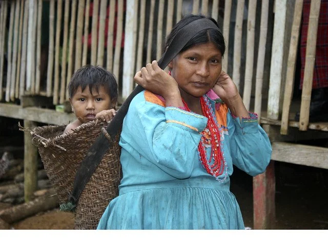 A young Embera mother carries her child in a handwoven basket pack, secured by a broad strap across her forehead. This traditional carrying method is common among Indigenous peoples of Panama and Colombia, where practical designs are rooted in centur
