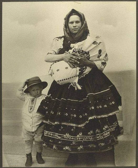 Dressed in elaborate embroidered layers, this woman stands with proud composure, holding her swaddled infant high against her chest. The baby is wrapped tightly in a cloth bundle, likely tied close beneath the mother’s shawl. Beside her, a young boy