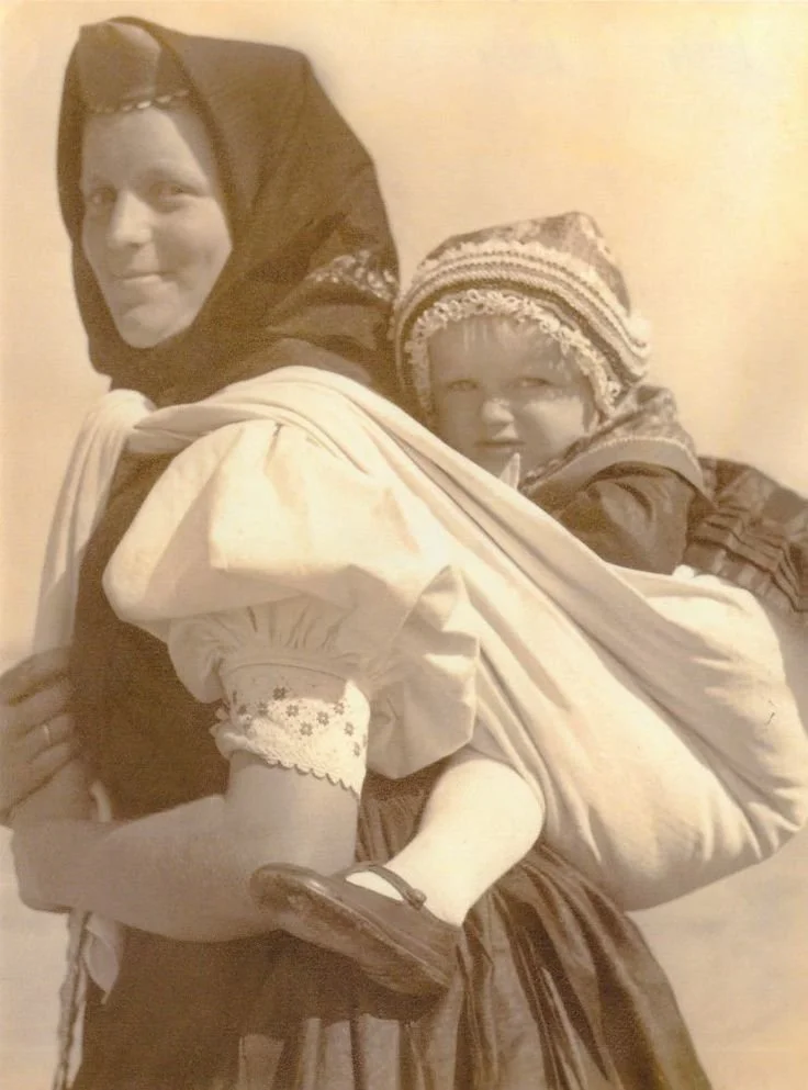 A mother smiles softly, her body turned just enough to reveal the toddler perched securely on her back. The child is wrapped in a long cloth, held high and snug against the mother’s torso with legs straddling her hips — an instinctive high back carry