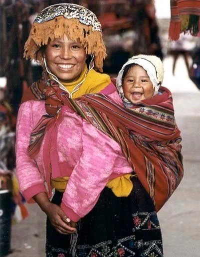 A mother smiles broadly as she walks through a vibrant market, her pink jumper vivid against the rich backdrop of textiles and stalls. On her back, her baby laughs with delight, eyes crinkled and mouth open wide in a joyful grin. The child is nestled