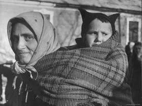 A woman carries a child wrapped securely in a large checked woollen shawl, worn over one shoulder and across her back in a traditional style common throughout Eastern Europe. Her headscarf and clothing suggest a rural setting, likely in the Carpathia