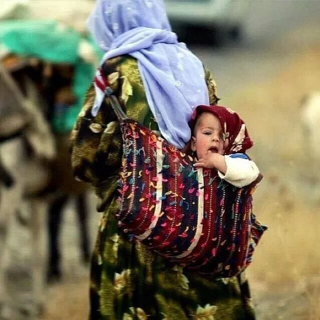 This vibrant image captures a traditional babywearing practice still alive in parts of Central and West Asia. A woman, dressed in richly patterned clothing and a lilac headscarf, carries a baby on her back using a wide, colourful woven cloth tied sec