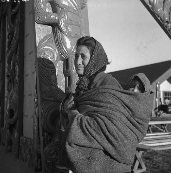 A Māori woman stands beside a carved wharenui post, holding her baby close in a soft woollen wrap. The fabric is carefully layered and tucked, forming a secure cocoon around the sleeping infant on her back — head covered against the cold, nestled int