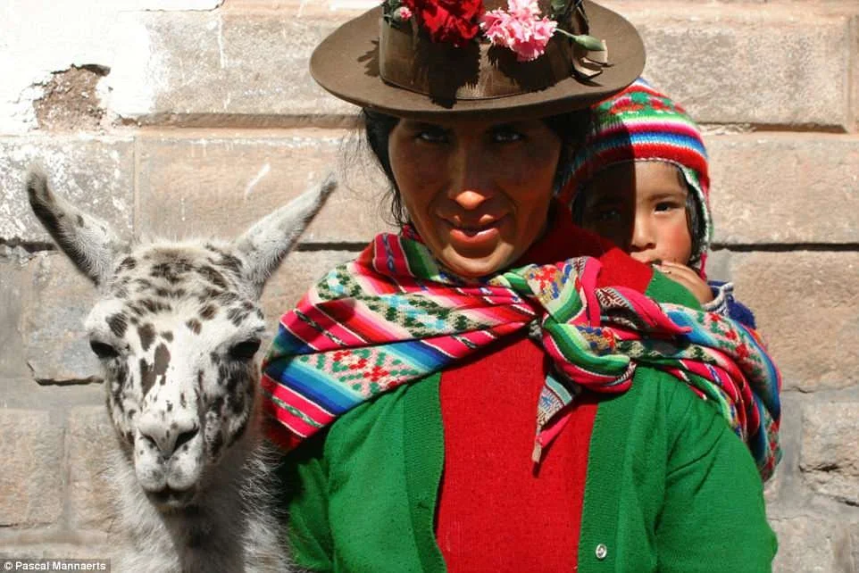 A Quechua mother stands proudly before a stone wall, her gaze warm and steady beneath the brim of her brown felt hat, which is adorned with bright carnations. She wears the rich greens and reds of traditional Andean clothing, layered for both functio