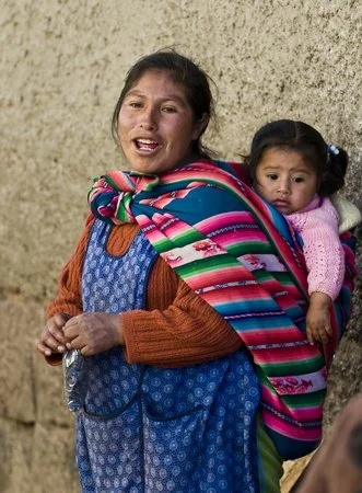 A mother pauses to speak beside a sun-warmed wall in Cusco, her child nestled close against her back. The little girl peeks out from a brightly woven manta, resting comfortably and securely as the day unfolds around them. Wrapped in vivid stripes of
