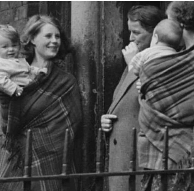 Two women stand at the edge of a city doorway, babies wrapped close in woollen shawls as they chat with a man in a long coat. The setting is unmistakably urban — iron railings in the foreground, brickwork and pillars behind them. The women’s faces ar
