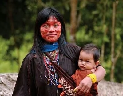 A young woman of the Achuar people stands calmly, her child held close in a simple front carry. She wears traditional face paint in deep red ochre, with lines drawn in symmetrical patterns that reflect both identity and ceremony. Beaded strands drape