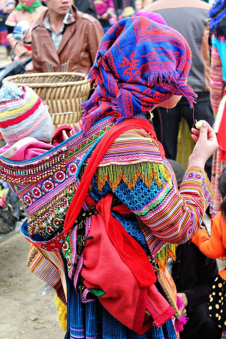 In a swirl of vivid colour and intricate detail, a woman walks through a bustling market, carrying her baby high on her back. Her carrier — a richly embroidered, structured cloth panel — is adorned with beadwork, floral patterns, and rows of tiny mir