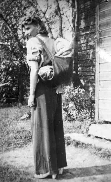 In this sunlit moment captured outside a wooden farmhouse, a Scandinavian woman carries her baby in a krip — a soft-bodied carrier sewn from sturdy fabric, worn high on the back and supported by wide shoulder straps. Popular across parts of rural Swe