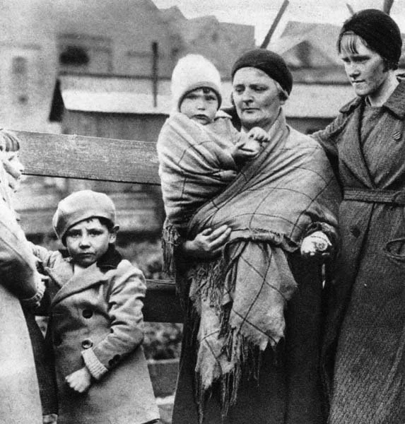 In this mid-20th-century photograph, a woman stands with quiet strength, her shawl drawn tightly across her chest in the traditional Welsh shawl carry. A young child, warmly dressed in a knit cap, is bundled high against her body, the patterned shawl
