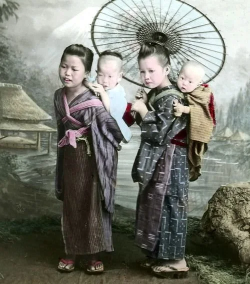 Two young girls stand quietly, each carrying a baby on their back, wrapped securely in cloth — an intimate portrait of sibling care and responsibility. Their posture is composed and steady, a testament to their familiarity with the task. Both girls w