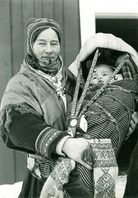 Wrapped in thick woollens and a richly adorned shawl, this Northern Sámi woman stands outside in the snow, her baby safely nestled in a beautifully decorated komsá cradleboard. Her pipe, the intricate silver jewelry, and the layered wool garments all