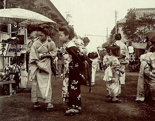 In a bustling street scene somewhere in early 20th-century Japan, women in kimono and traditional wooden geta gather in quiet conversation, surrounded by the gentle chaos of a summer festival or market day. Among them, several carry babies on their b