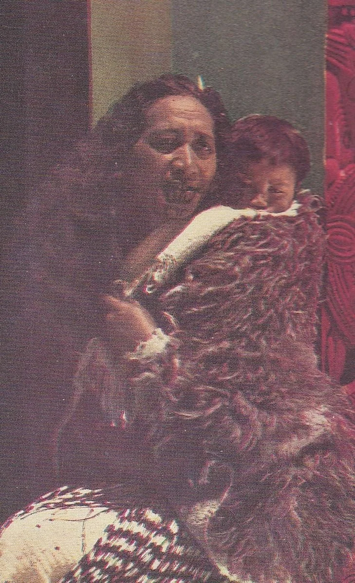 Wrapped in a richly textured korowai, this Māori mother carries her sleeping child high on her back, close to her heart. The image, possibly a hand-coloured postcard or print from the mid-20th century, shows the mother in traditional dress — her long
