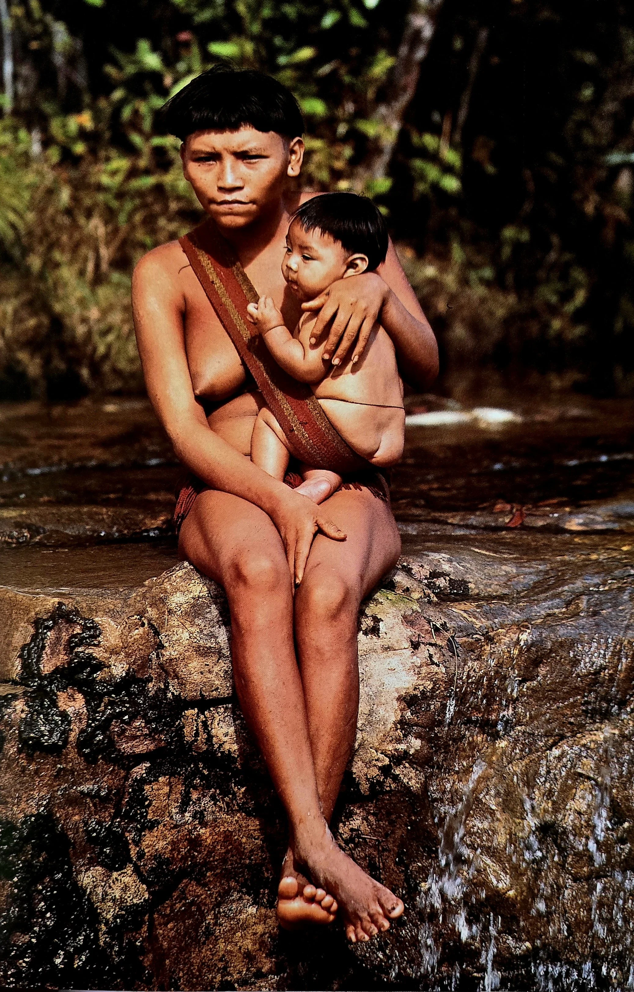 A young mother of the Yanomami people rests on a rock beside a river deep in the Amazon, her baby settled securely in a broad woven sash that supports their weight and keeps them close. The sling crosses diagonally across her torso, anchoring the bab