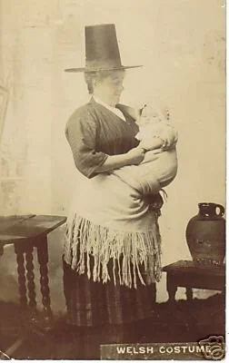 Dressed in traditional Welsh costume — including the iconic stovepipe hat and fringed shawl — this woman cradles her baby securely, wrapped and held close in a wide piece of cloth. Though taken in a studio setting, likely as a posed postcard, the ima