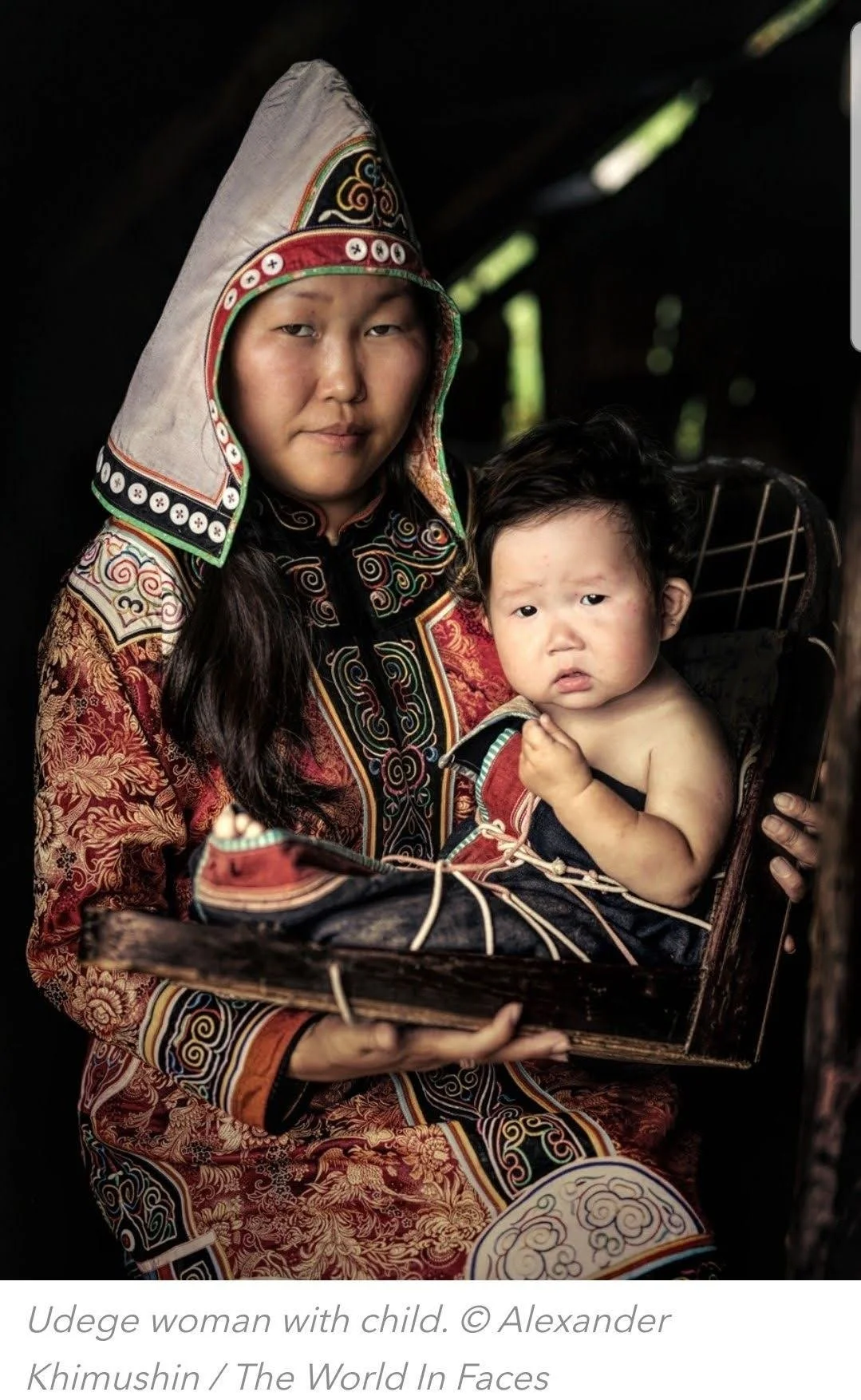 In this striking contemporary portrait, an Udege woman wears traditional dress richly embroidered in red, gold, and forest green, her tall headdress edged with protective motifs. Her baby rests securely in a wooden cradleboard, swaddled and fastened
