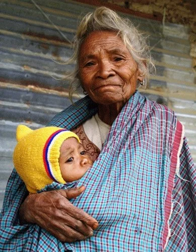 An elder woman holds a baby close to her chest, wrapped together in a wide checked cloth that crosses her body like a shawl. Her weathered hands and quiet expression speak of deep experience and enduring care. The infant is nestled upright against he