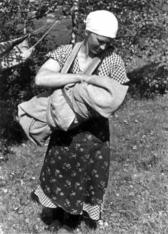 On a sun-dappled hillside beneath the birch trees, a Scandinavian mother smiles as she adjusts her baby, securely wrapped and nestled in a traditional fatle — a cloth baby carrier once widely used across Norway, Sweden, and parts of Finland. The padd