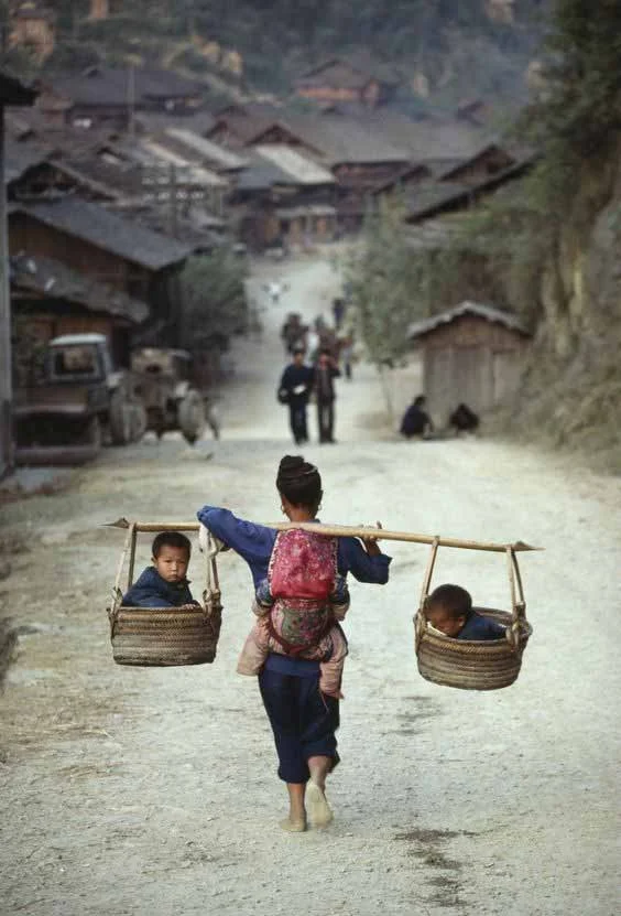 A barefoot mother walks along a dirt road through a rural Chinese village, framed by weathered wooden houses and steep forested hills. She carries three children at once — one tucked securely into a soft cloth carrier on her back, and two nestled in