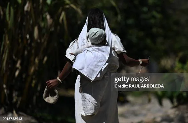 A Kogi mother walks a sunlit mountain path, her baby tied snugly to her back with soft white cloth. Her gait is steady, her arms free to carry the tools and threads of daily life—an age-old balance between nurture and necessity. The baby’s head rests