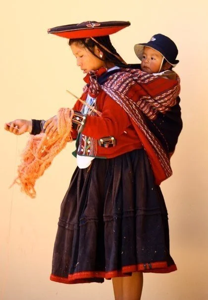 A Quechua mother from the Peruvian Andes carries her child in a traditional lliqlla, a woven mantle folded and tied across her chest. The baby rests high and secure against her back, upright and close, in the protective curve of cloth and connection.