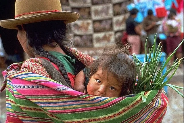 Amid the colourful bustle of a highland market, a child rests quietly in the woven sling of their mother’s aguayo. The striped cloth, alive with vivid colour and pattern, is wrapped securely around the woman’s shoulders and back, forming both carrier