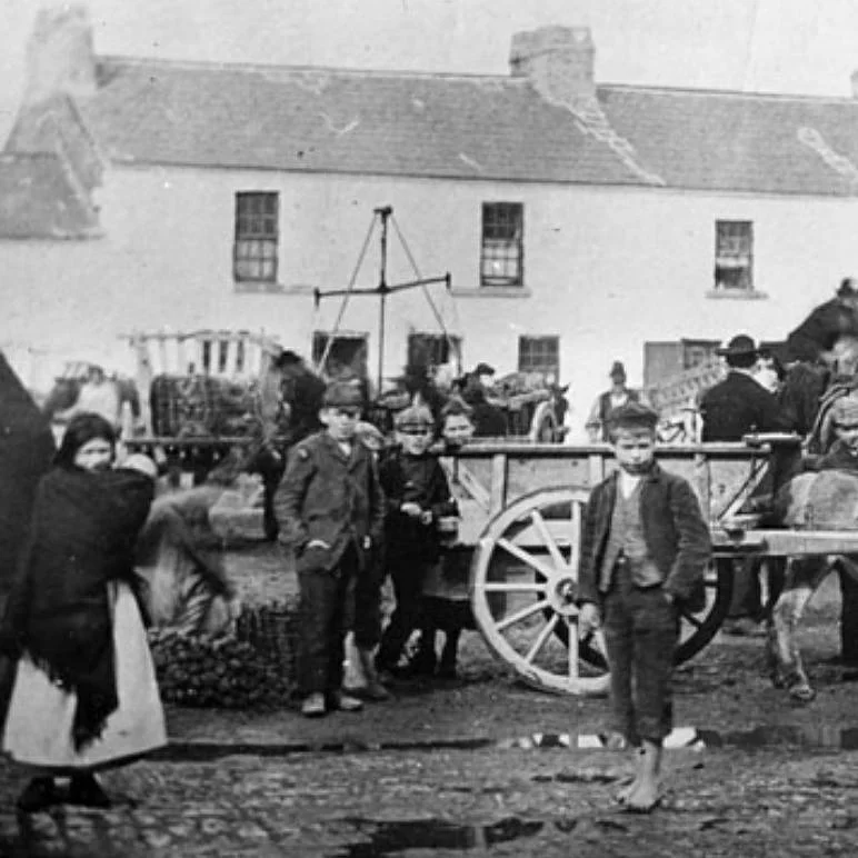 In a busy Irish market square, children cluster near a horse-drawn cart while farmers unload goods in the background. The cobblestone ground is scattered with puddles, baskets, and produce. Behind them, a row of whitewashed buildings lines the street