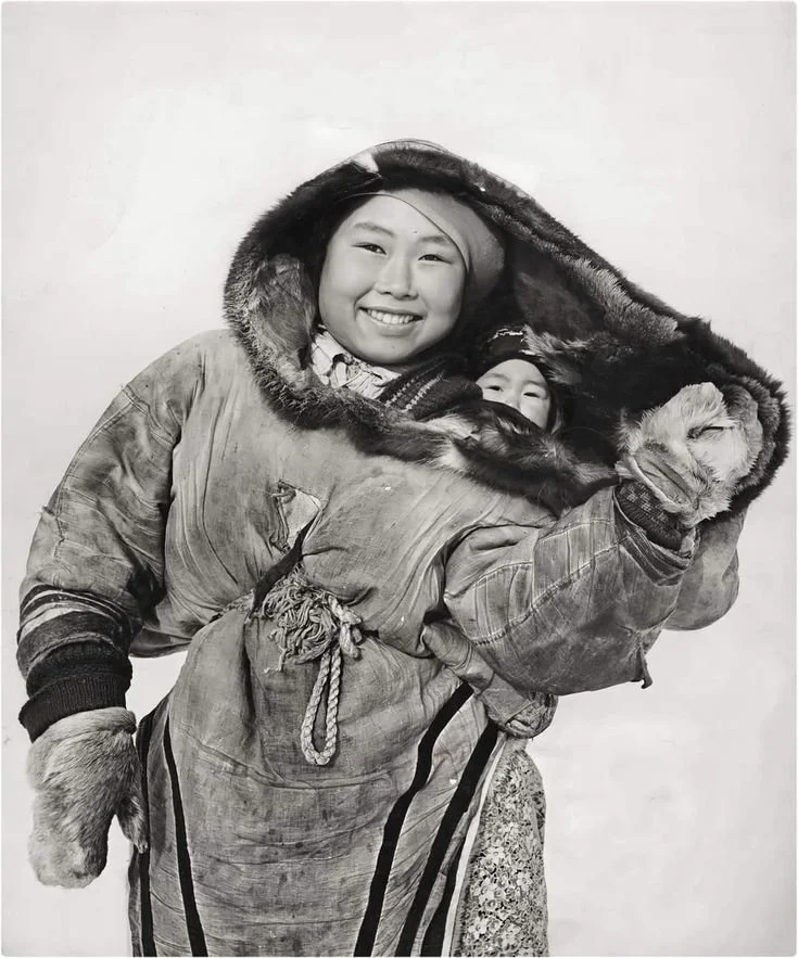 With a joyful smile and arms lifted in mid-gesture, this Inuit mother proudly wears her traditional amauti, the iconic fur parka uniquely designed for carrying babies in the Arctic. Her child is nestled high between her shoulders, peeking out from th