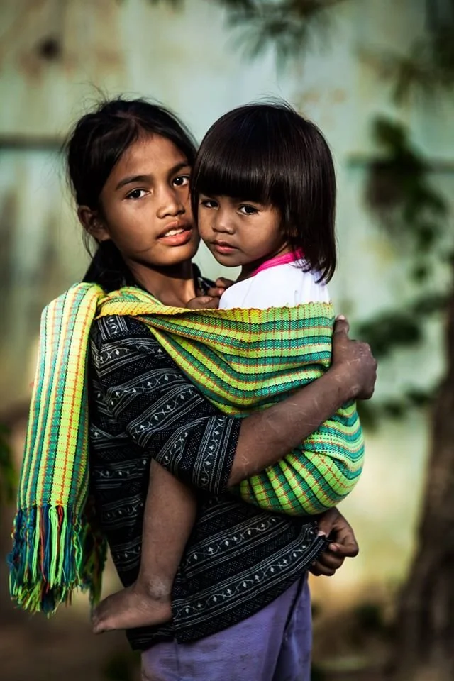 A young girl gazes steadily at the camera as she carries a slightly younger child in a brightly coloured, handwoven rebozo-style cloth. The fabric, wrapped securely over one shoulder and across her torso, cradles the child on her hip with practiced e