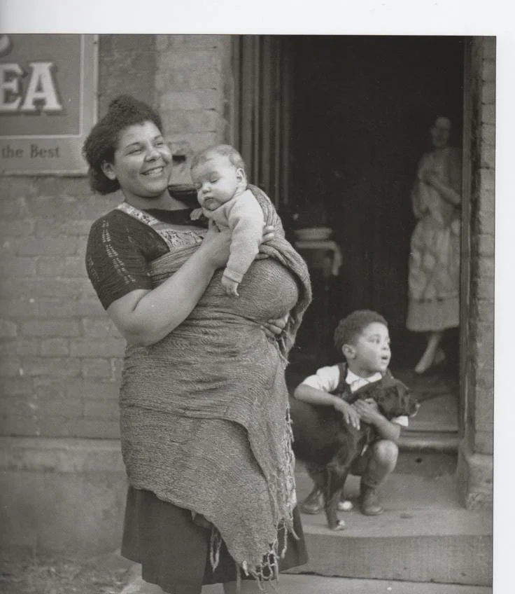 A Welsh mother stands smiling in the street, wrapped in a woven shawl that holds her sleeping baby close. The wide cloth crosses her chest and loops over her shoulder in the traditional Welsh shawl carry — a method passed down through generations, kn