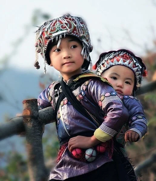 A young girl from one of the ethnic minority communities in the mountains of southwest China—possibly Miao or Hmong—carries her sibling nestled securely on her back. Their finely embroidered jackets and richly adorned headdresses reflect deep cultura