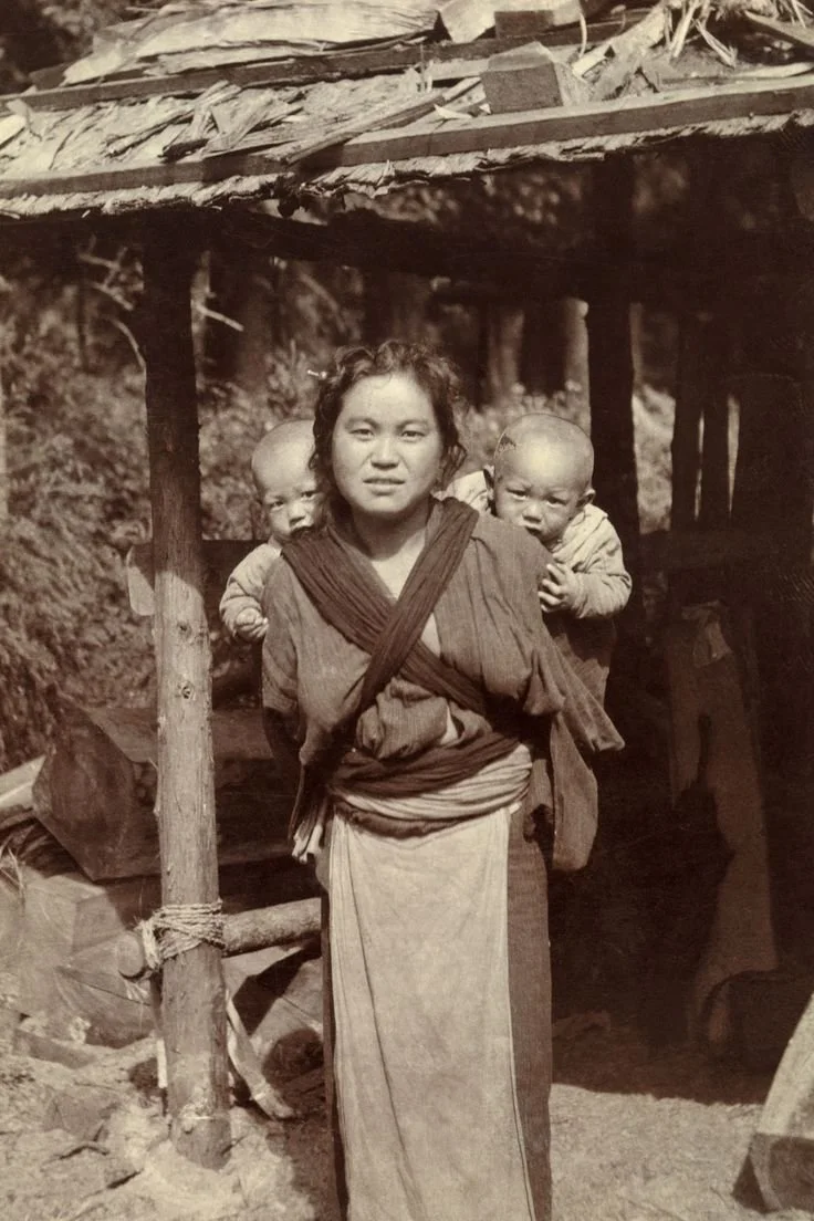 A mother stands beneath a timber shelter, two babies tucked securely against her back. She wears a long rectangular cloth, crossed high across her chest and tied firm around the waist — a style widely used across rural China, where caregivers have lo