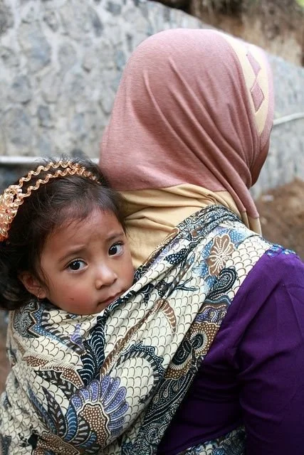 In this tender photograph, an Indonesian mother carries her child on her back using a traditional selendang, a long rectangular piece of batik fabric. The richly patterned cloth, featuring classic Indonesian motifs, is wrapped securely around the chi