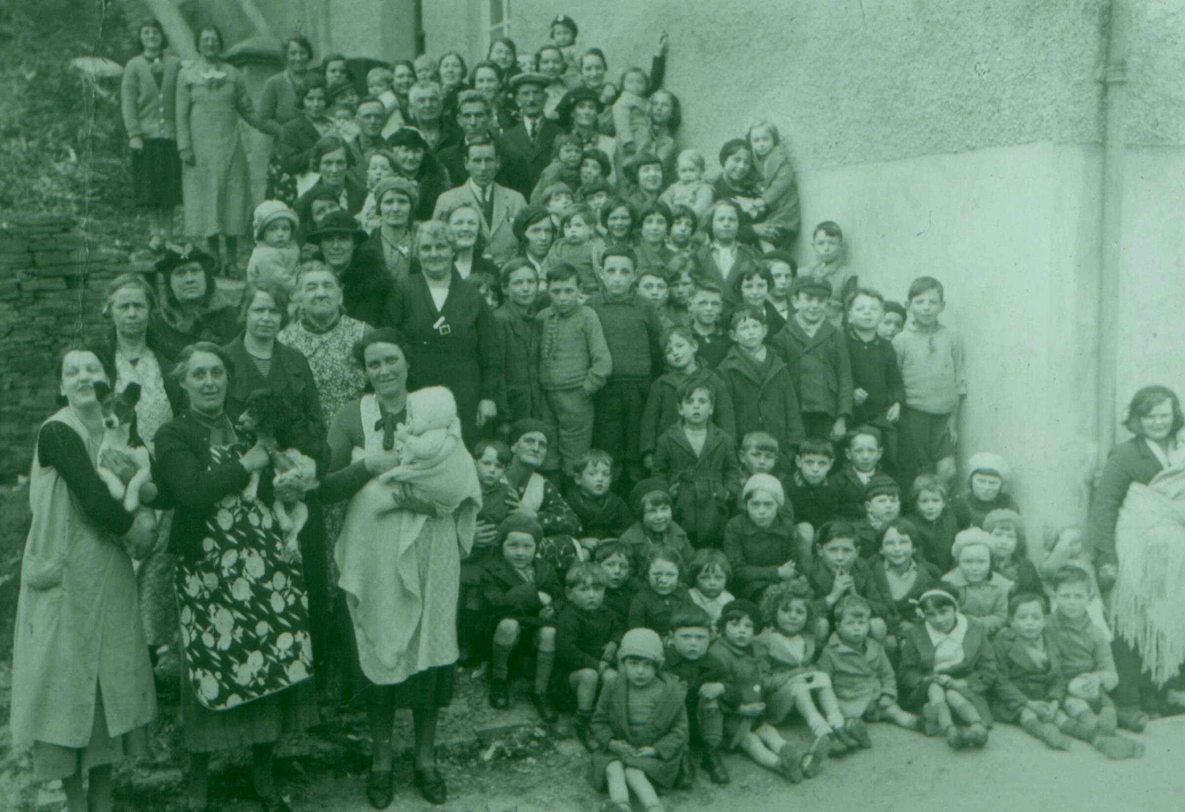 A sea of faces fills the frame — women, children, elders, and even dogs — gathered tightly together outside a building that may have been a school, parish hall, or community centre. The occasion is unknown, but the sense of solidarity is unmistakable