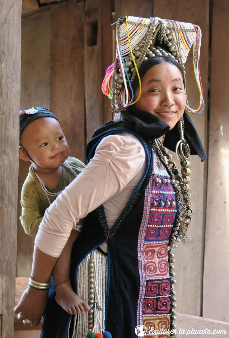 A mother from the Akha community smiles as she turns toward the camera, her child peeking out contentedly from a richly adorned back carrier. Her traditional headdress is unmistakable — a tall structure of silver coins, beads, and bright cords that s
