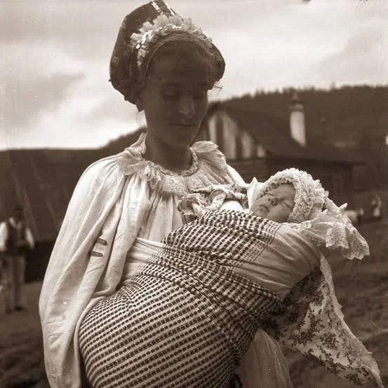 A young mother gazes tenderly at her swaddled infant, who is bound snugly in a gingham cloth and secured in a side carry against her hip. The baby wears a delicate lace bonnet, while the mother’s embroidered blouse and floral headscarf suggest region