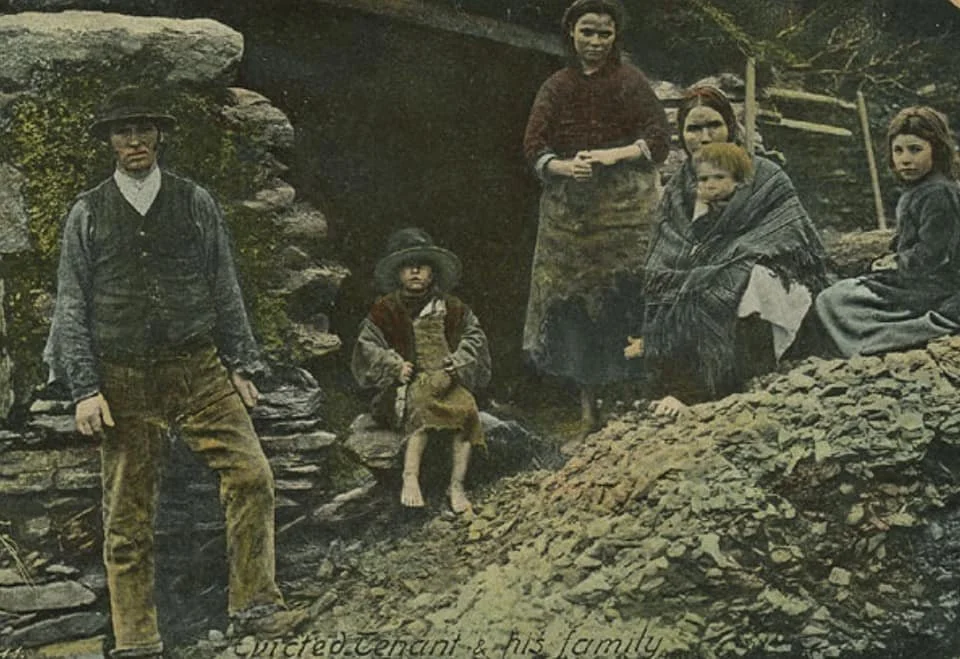 A family stands on uneven ground outside their ruined cottage, the stonework behind them crumbling and overgrown. The handwritten caption — “Evicted Tenant & his family” — speaks plainly to the hardship unfolding.
The mother, barefoot, wraps her chi