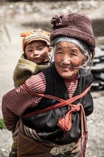 An elderly Tibetan woman carries a young child on her back in a rural mountain village. Her deeply lined face is full of warmth and resilience, and she smiles gently at the camera. The baby is securely bundled in a thick, earth-toned cloth wrap that