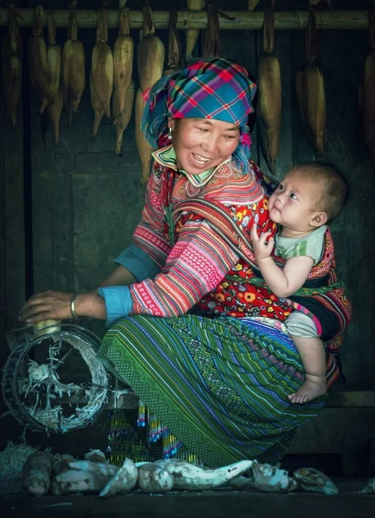 A mother works beside a hand-cranked shredder, preparing food while her baby nestles safely on her back in a vibrant carrier stitched with bold florals. Her skirt and headscarf, rich in layered patterns and colour, mark her as a member of the Flower