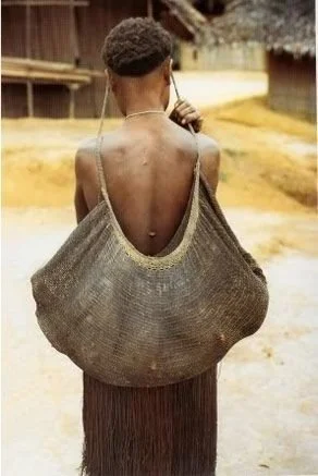 In this striking image, a woman from Papua New Guinea carries her baby in a traditional bilum, a handwoven string bag that is deeply woven into the cultural fabric of the region. The bilum is worn over her head and rests along her back, creating a ha