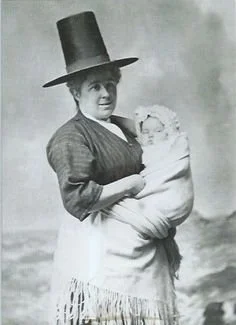 A Welsh woman stands posed before a painted backdrop, her tall stovepipe hat and fringed shawl unmistakably marking her cultural identity. She holds a swaddled baby at her front, snugly wrapped in her large woollen shawl — a time-honoured Welsh tradi