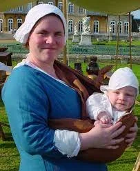 A woman in historical dress stands confidently, smiling as she carries her baby in a simple cloth sling across her front. The child is upright, well-supported, and content, dressed in a white bonnet and gown that matches the mother's linen head cover